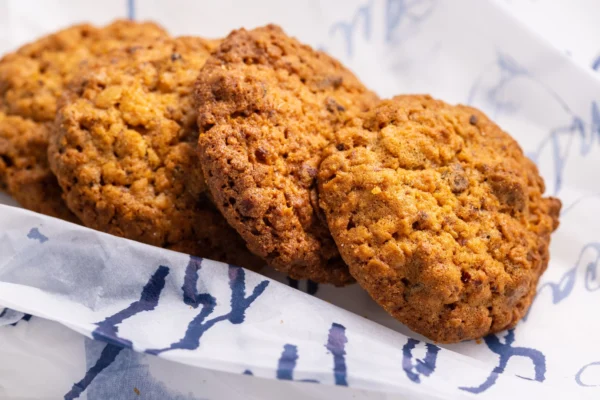 Guilherminas da Fábrica Santo António, close-up de biscoito dourado com pedaços de amêndoa e aveia, sobre papel seda estampado